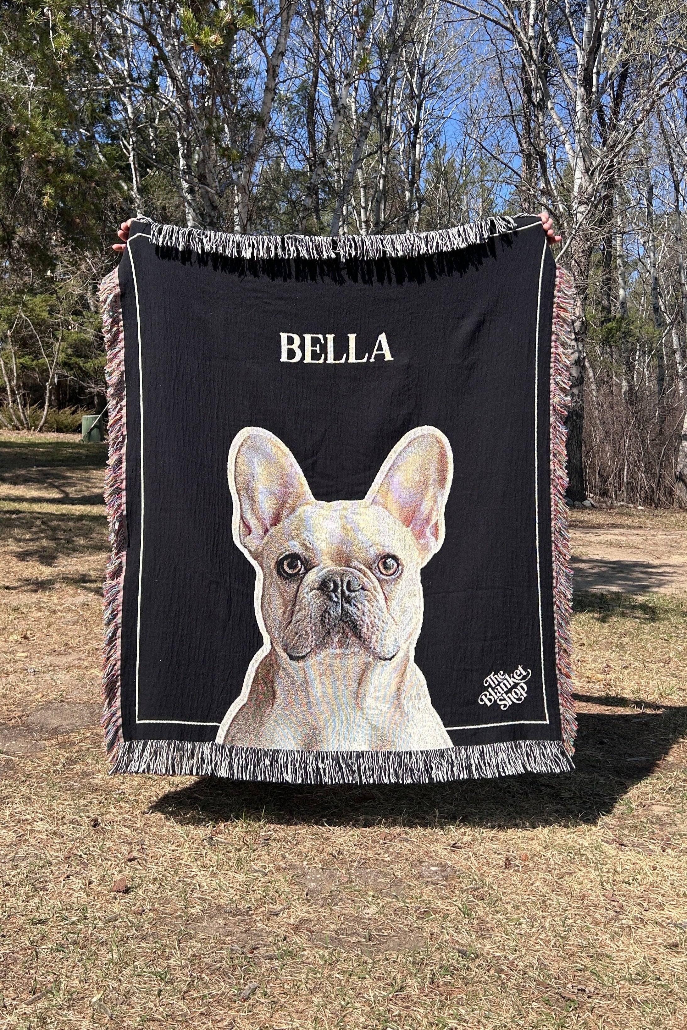 Custom throw blanket with a dog illustration and 'BELLA' text, held up outdoors with the Blanket Shop Logo