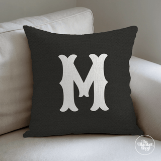 Black pillow with a white letter 'M' on a beige couch with The Blanket Shop Logo