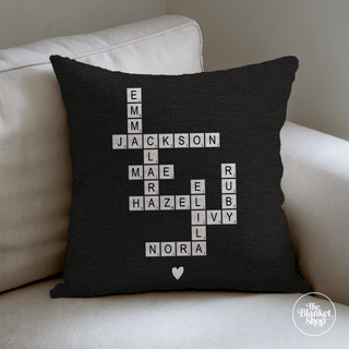 Black pillow with white crossword-style names on a beige couch with The Blanket Shop Logo