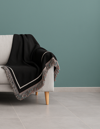 Corner of custom black throw blanket with colorful fringe on a white couch with a teal background wall from The Blanket Shop