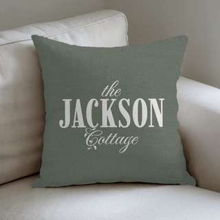 Green pillow with 'the JACKSON Cottage' text on a beige couch and The Blanket Shop Logo