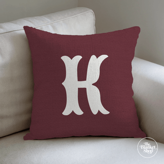 Maroon pillow with a white letter 'H' on a beige couch and The Blanket Shop Logo