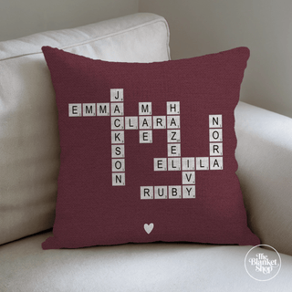 Maroon pillow with white crossword-style names on a beige couch with The Blanket Shop Logo