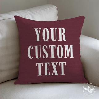 Red pillow with 'Your Custom Text' on a beige couch, brand 'The Blanket Shop' visible.

