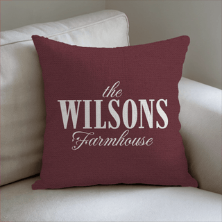 Red pillow with 'the WILSONS Farmhouse' text on a beige couch with The Blanket Shop Logo