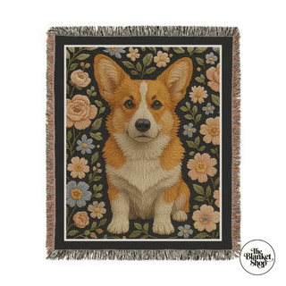 Woven blanket with a corgi design surrounded by flowers on a black background, featuring 'The Blanket Shop' logo.