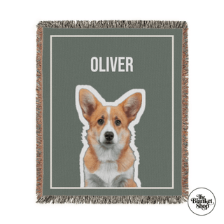 Woven blanket with a corgi illustration and 'OLIVER' text on a green background, featuring 'The Blanket Shop' logo.

